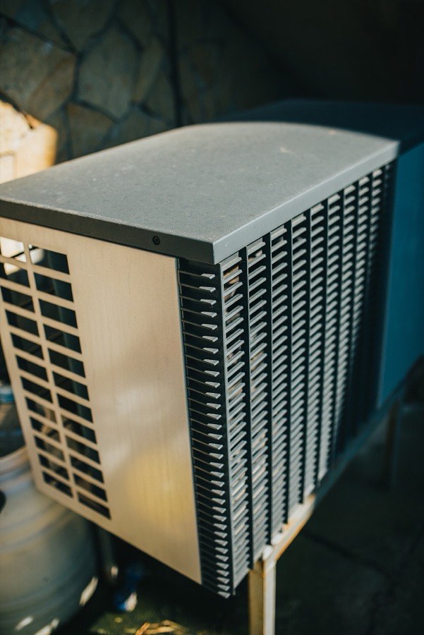 The outdoor condenser of a central air conditioner standing beside a house, its coil fins exposed along the side of the cabinet